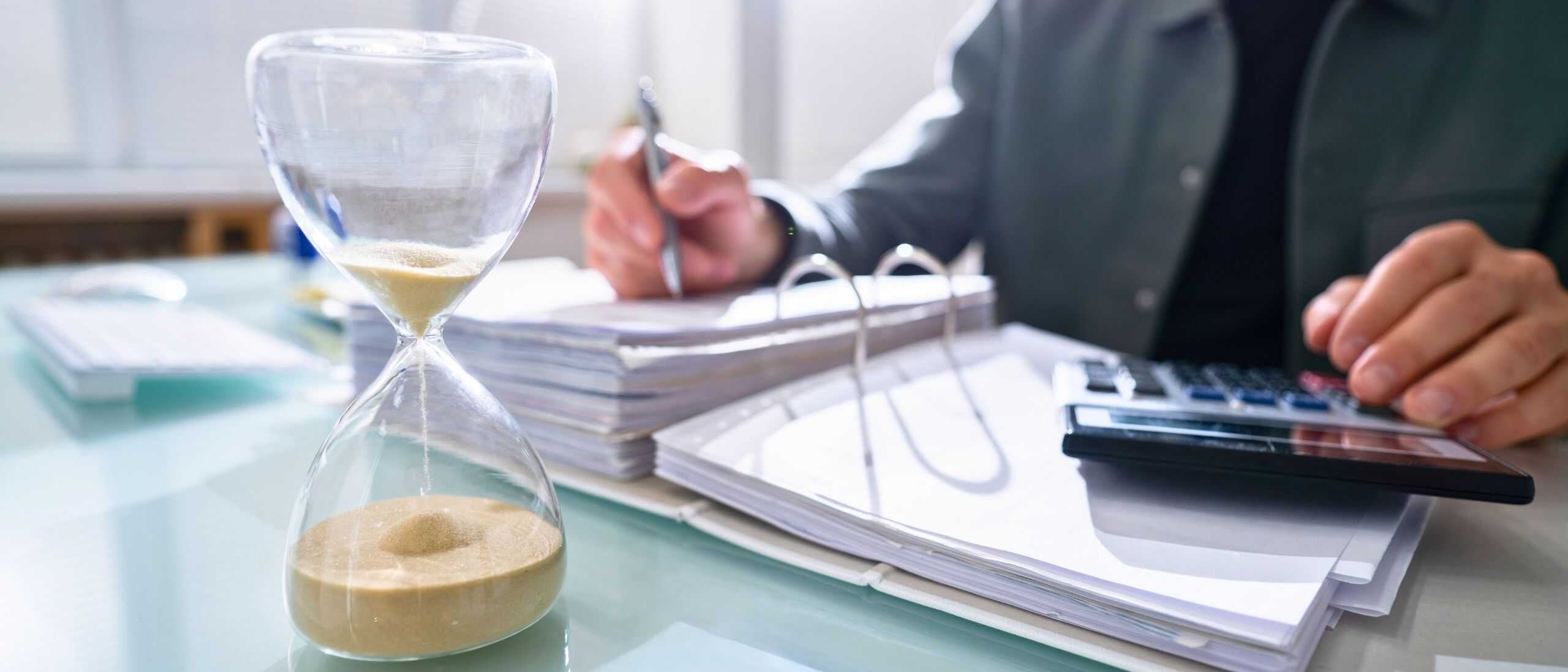 Stundenleitende Sanduhr und Arbeitstisch mit Dokumenten, Stift, Taschenrechner und Person im Anzug, die komplexe Unternehmensarbeit in einem modernen Büro erledigt.